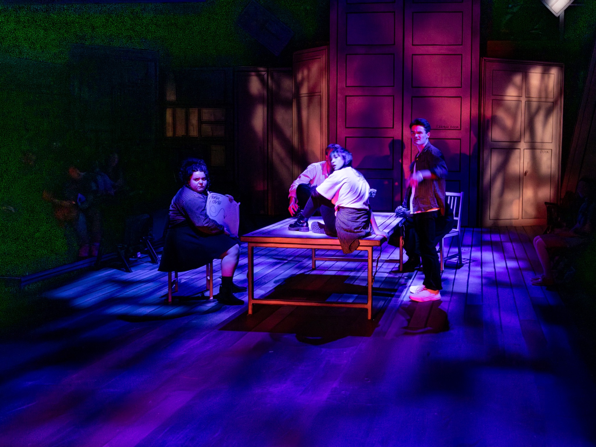Performers gather around a table in blue and magenta light.